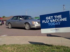 Yard sign reads "GET THE FLU VACCINE TODAY" with cars lining up for the drive-thru flu shot event.
