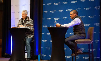 CEOs of Sanford Health and Intermountain Healthcare sit at podiums on a stage for a press conference. Blue and white backdrops show the logos of both organizations as they announce their merger.