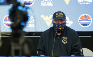 Bob Huggins talking with the media during a Zoom call on Tuesday. He is wearing a mask and hat with WVU logos and sitting in front of a screen with the Crossover Classic logos.