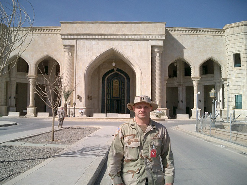 A Sanford Health physician in Army camouflage stands in the road in front of a grand palace in Iraq.