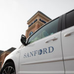 A Sanford Health minivan courier is parked outside a clinic with a brick tower.