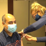 A Sanford Health doctor, seated, gets a COVID-19 shot in his arm from a colleague.
