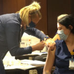 A Sanford Health nurse leans over a female colleage to give her a COVID-19 shot in her left arm.