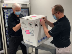Two men in surgical masks and black shirts lift a white shipping box of COVID-19 vaccine off a cart.