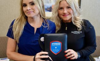 Two Sanford Health nurses face the camera while holding their invention, a black pad with a blue shield that says "Sternum Protect."
