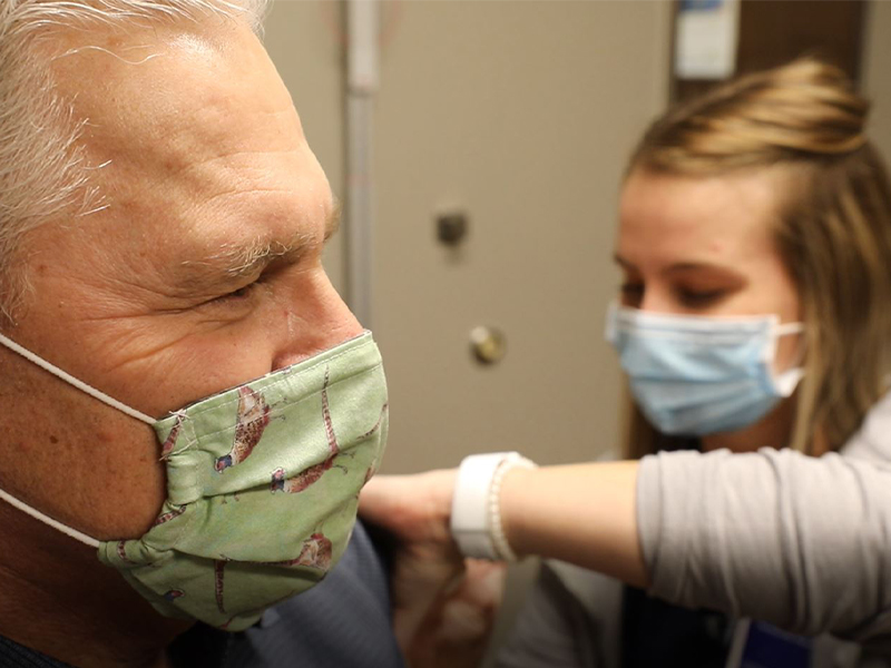 Randy Bury, masked and in profile, gets a shot in the arm from a nurse in an exam room.