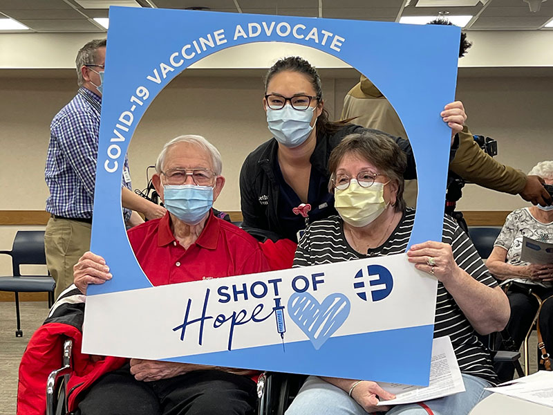 A Sanford Health nurse poses with two older adult patients with a poster frame that says "COVID-19 vaccine advocate: A shot of hope"