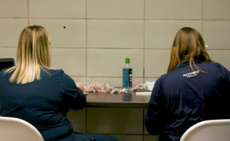 Two female Sanford Health workers, seen from behind, sit at a table preparing COVID-19 vaccine in syringes.