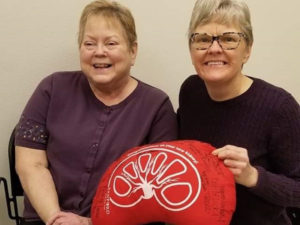 Two mature women smile and hold a red kidney-shaped pillow.