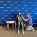 Sanford Health nurse gives vaccine in teenage girl's arm against Sanford Health logo backdrop.