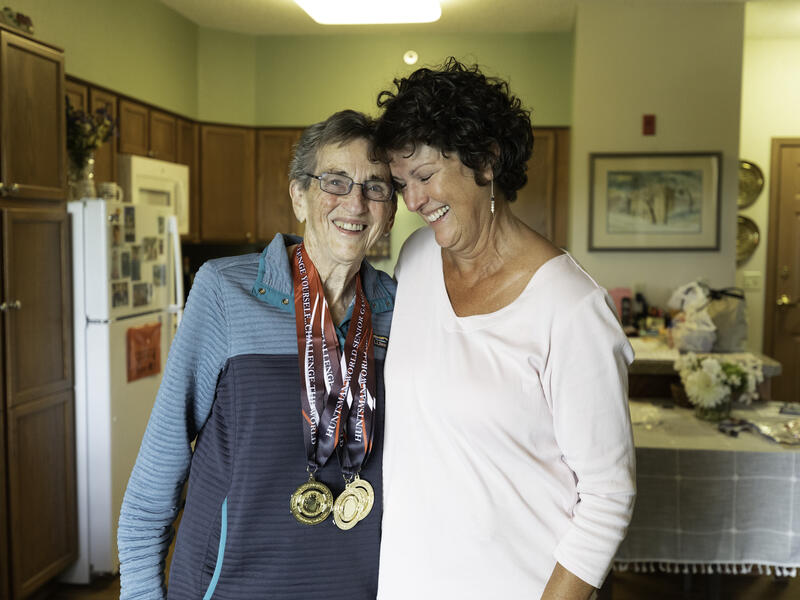 A senior woman with gold medals around her neck hugs her adult daughter in her kitchen at Good Samaritan Society.