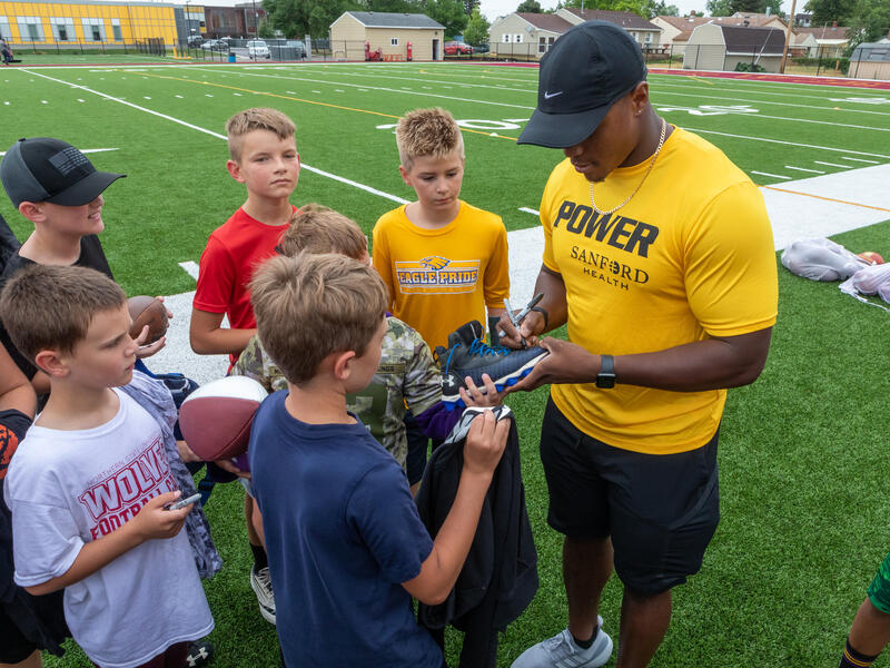 CJ Ham, in gold Sanford POWER shirt, signs autographs for young football players at camp.