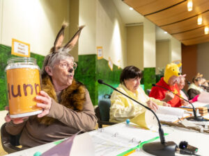 Older adults in costumes of Rabbit, Christopher Robin, Winnie the Pooh and others read from microphones while sitting behind a table. Rabbit is holding a giant pot labeled "hunny."
