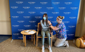 Sanford Health nurse gives vaccine in teenage girl's arm against Sanford Health logo backdrop.