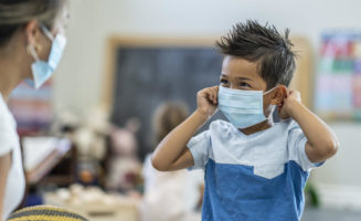 Cute little boy putting on a protective face mask while in daycare during phase two of reopening after the COVID-19 outbreak.