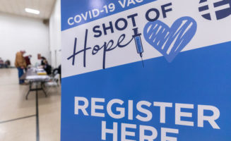 Blue and white poster in a gym reads "COVID-19 VACCINE. A SHOT OF HOPE. REGISTER HERE." People at a registration table in the background.