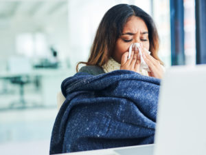 A young businesswoman blowing her nose with a tissue and covering with a blanket in a modern office.