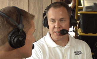 A Sanford Health patient talks to an unidentifiable man in the foreground while wearing a headset. They are sitting in the press box at a football game. His polo shirt says "Midco Sports."