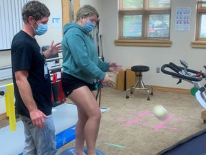 In a clinical gym, a young woman throws a ball to the floor while balancing on one foot. Her Sanford Health physical therapist stands next to her.
