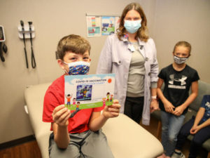 A 10-year-old boy displays his COVID-19 vaccine sticker chart showing he has one more dose to go. His mom, who is a doctor, and sister and brother are nearby in the exam room.