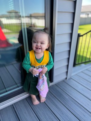 Laughing little girl in a Green Bay Packers cheerleader outfit stands barefoot on a deck against a sliding glass door.