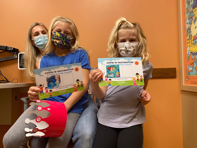 Two little girls share their mom's lap and display their COVID vaccination cards in an exam room.