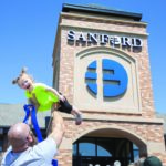 A father lifts his giggling toddler daughter high in the air outside a Sanford Health clinic grand opening.