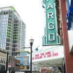 The iconic Fargo Theater sign stands out in teal blue against a cloudy sky. Its marquee reads in red: "WASH YOUR HANDS AND BE EXCELLENT TO EACH OTHER."