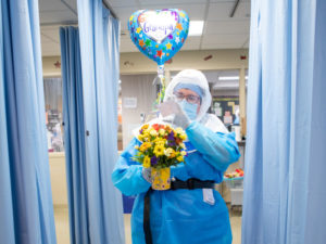 A Sanford Health COVID unit nurse in full PPE carries flowers and a heart-shaped balloon that says "Get Well Grandpa."