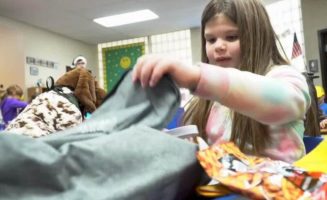 A girl unpacks a backpack on her school desk, including a stuffed dog and bag of pretzels.