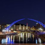 Nighttime view of the Arc of Dreams sculpture lit up in blue. Downtown Sioux Falls buildings in the background are reflected on the river.