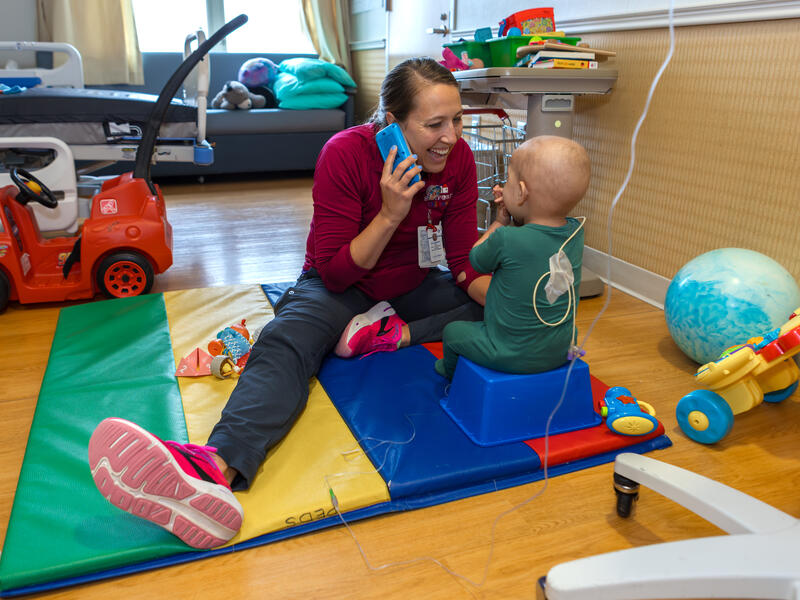 Hospital worker plays on the floor with a toddler patient, pretending to excitedly talk on the phone. The young patient has a tube attached to their back.