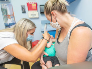 A Sanford Health APP (advanced practice provider) uses a scope to examine a baby, held in their mother's arms in an exam room.