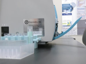 A FIT testing machine labeled "Polymedco, Inc." sits open on a countertop with trays of test tubes next to it.