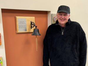 Sanford Health patient poses by a bell on the wall patients ring after chemo.