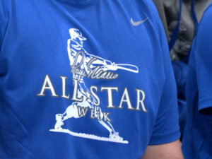 Closeup of blue shirt with white logo, a baseball player behind "Roger Maris All-Star Week"