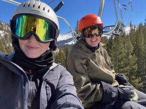 A couple in ski goggles takes a selfie while on a ski lift on a sunny day.
