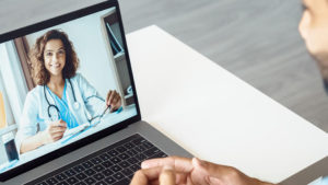 Doctor smiles from the screen of a laptop while patient in foreground talks to her.