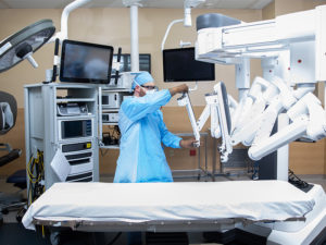 Sanford Health surgeon in scrubs adjusts the arm of a machine in the operating room. Above him are large screens for imaging.