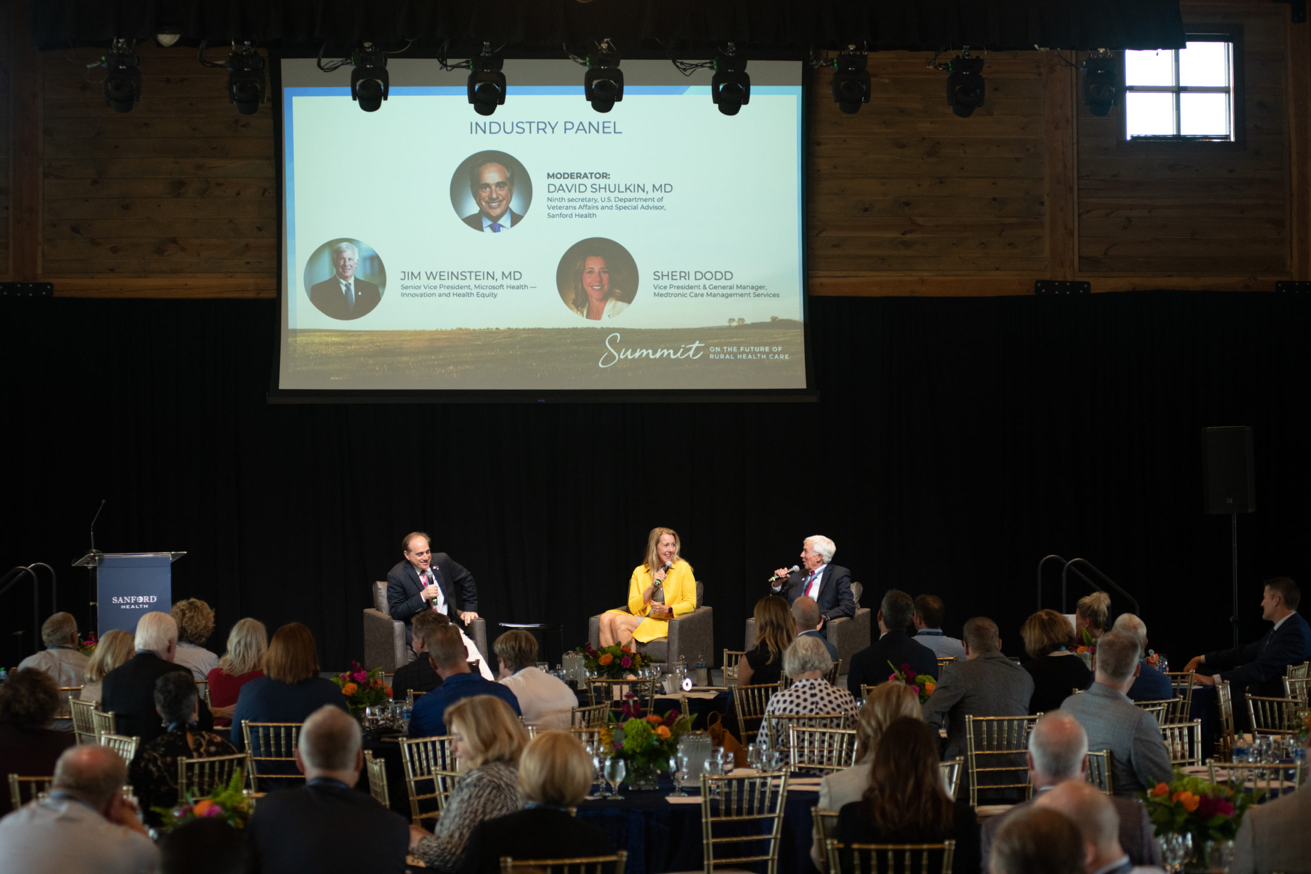 Three businesspeople sit onstage with their photos and titles on a screen above them as a crowd listens from tables in the foreground.
