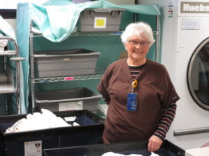Mature women in scrubs smiles among laundry shelves at a Sanford hospital.