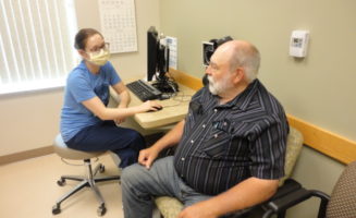 Nurse navigator in face mask sits down to meet with a cancer patient in an exam room.