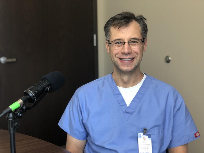 Dr. Chris Johansen smiles while wearing scrubs and sitting next to a microphone.