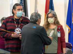 Two Sanford Health workers in surgical masks stand at the front of a flag-draped room, one holding a ceremonial pipe, while a Native American advisor touches them with an eagle feather.