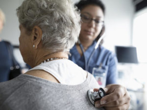 Female nurse using stethoscope on back of senior patient in clinic examination room.