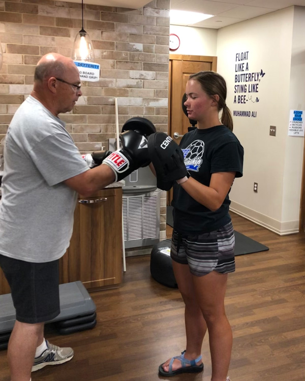 A mature man and young woman spar in a gym for Parkinson's patients. Words on the wall say "Float like a butterfly, sting like a bee. - Mohammad Ali."