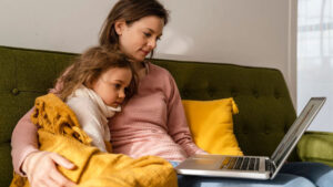 Mom snuggles daughter on couch during virtual care visit on her laptop.