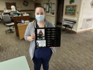 Nurse in a surgical mask holds up plaque of DAISY Award winners with her name and photo added.