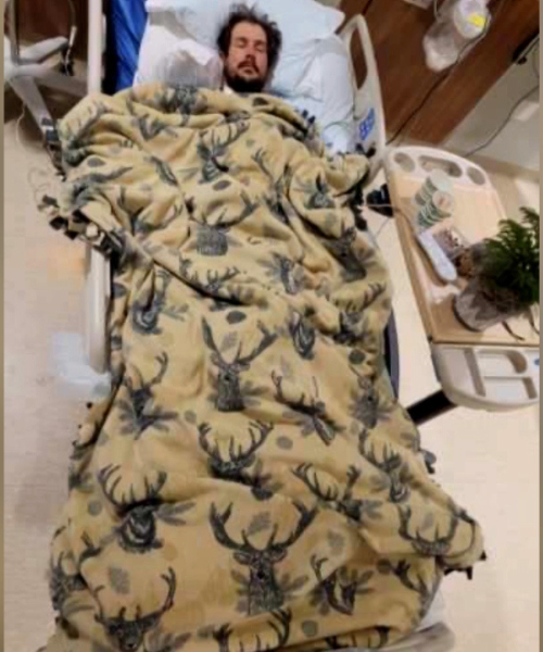 Mitchell Amundson lies in a hospital bed covered with a brown blanket in a deer head pattern.