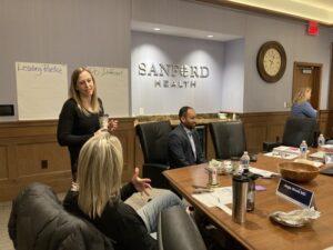 Sanford Health clinicians chat about clinician well-being over coffee in a conference room. Two large notes on the wall read "Leading Practice" and "Do Different."